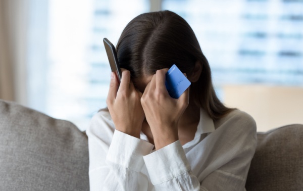 A woman holds her head with a phone in one hand and a bank card in the other.